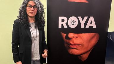Eine Frau mit langem lockigen und graumeliertem Haar steht neben einem Filmplakat, das ein Frauengesicht mit einer breiten schwarzen Augenbinde zeigt, auf der Roya in Weiß geschrieben steht.