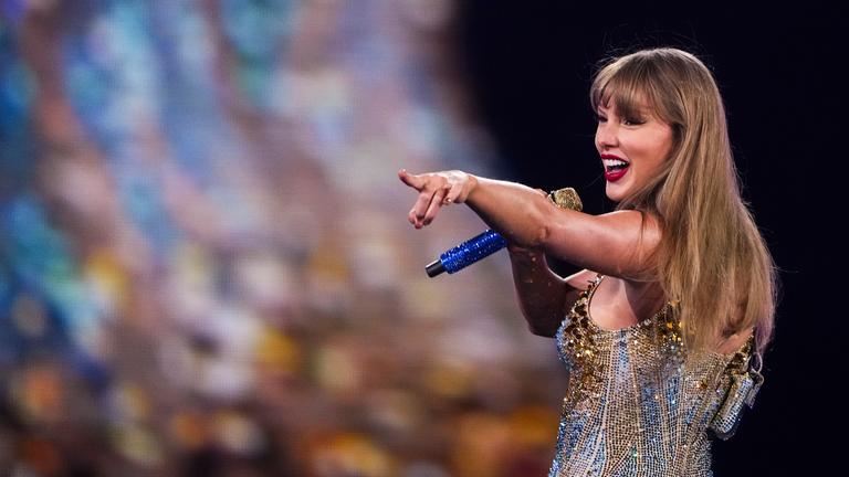Taylor Swift im Glitzer-Kostüm dreht sich nach hinten um, lächelt und zeigt mit augestrecktem Arm von sich weg. In der Hand hält sie ein glitzerndes Mikrofon.