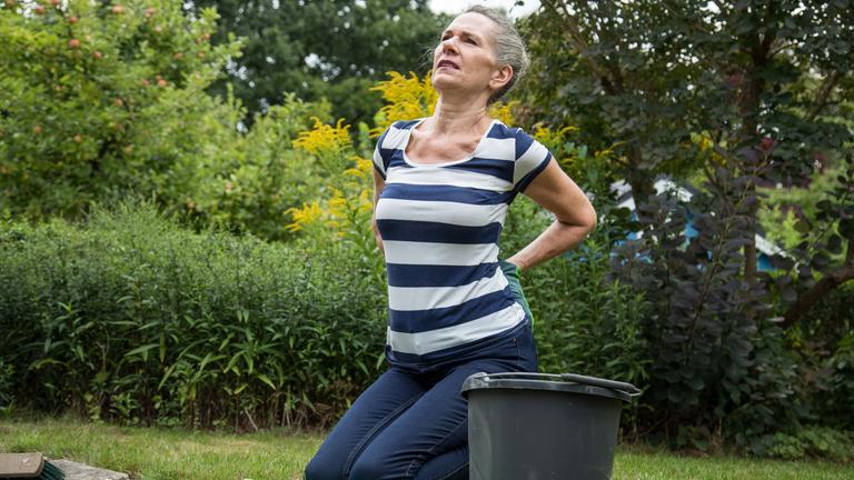 Eine Frau kniet beim Gärtnern auf dem Boden und hält sich den unteren Rücken, der ihr Schmerzen bereitet. 