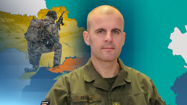 Oberst Reissner in der Mitte. Im Hintergrund sind Kartenumrisse und ein Soldat mit Waffe zu sehen