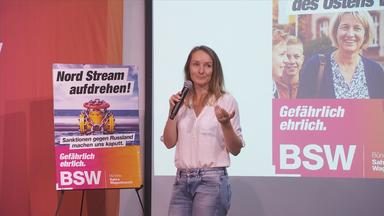 Eine Frau steht vor einer Leinwand neben ihr ist ein Plakat zu erkennen vom BSW, ein weiteres ist auf der Leinwand abgebildet 