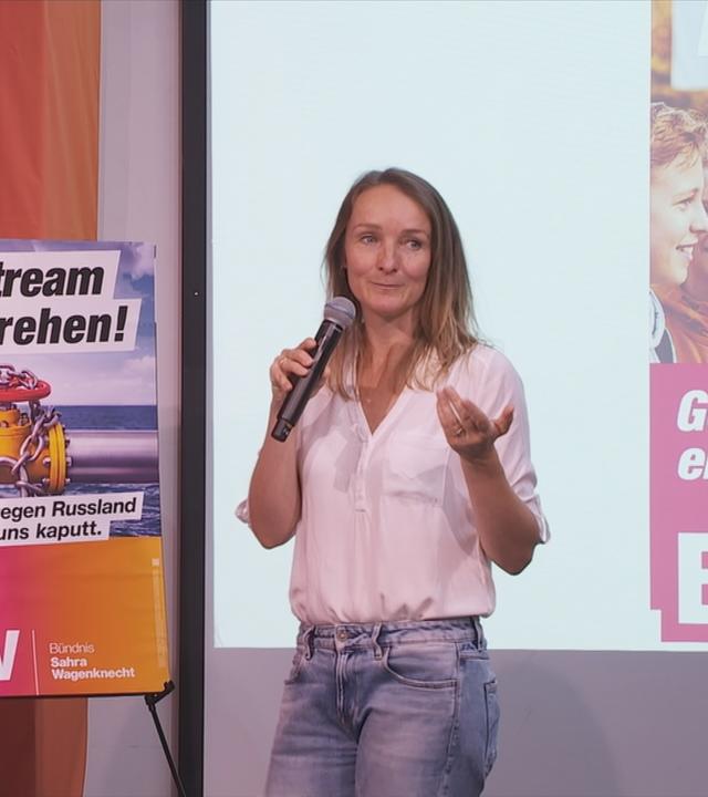 Eine Frau steht vor einer Leinwand neben ihr ist ein Plakat zu erkennen vom BSW, ein weiteres ist auf der Leinwand abgebildet 