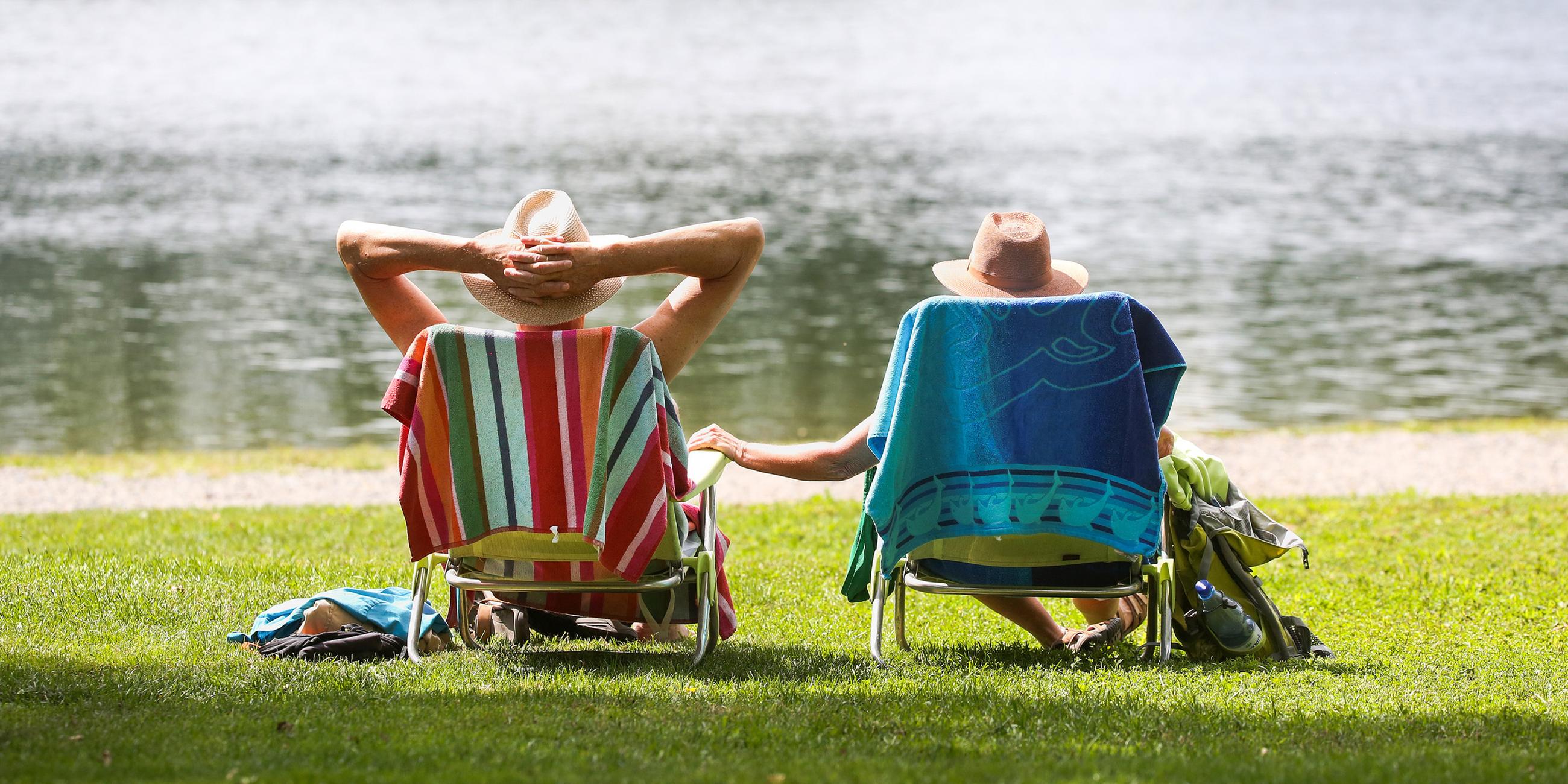 Ein Mann und eine Frau mit Strohhüten sitzen an einem Badesee in ihren Stühlen in der Sonne. (Archiv)
