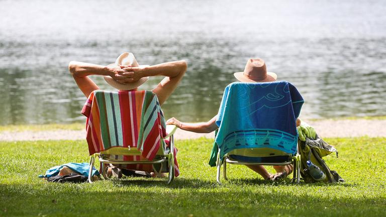 Ein Mann und eine Frau mit Strohhüten sitzen an einem Badesee in ihren Stühlen in der Sonne. (Archiv)