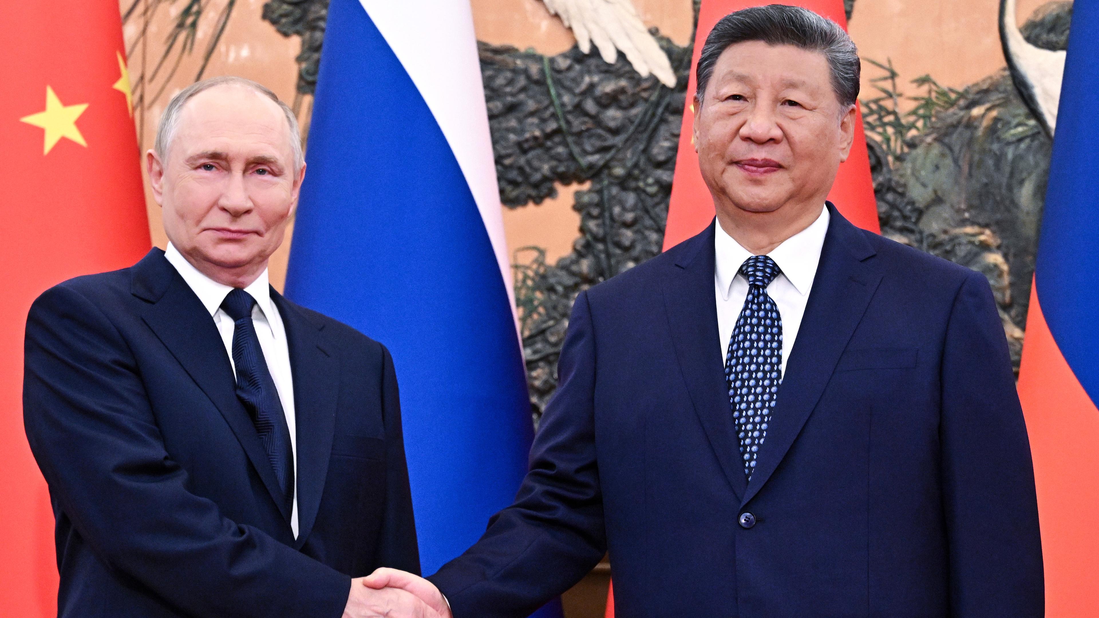 Der russische Präsident Putin und der chinesische Präsident Xi, wie sie sich die Hände schütteln vor den Flaggen von Russland und China