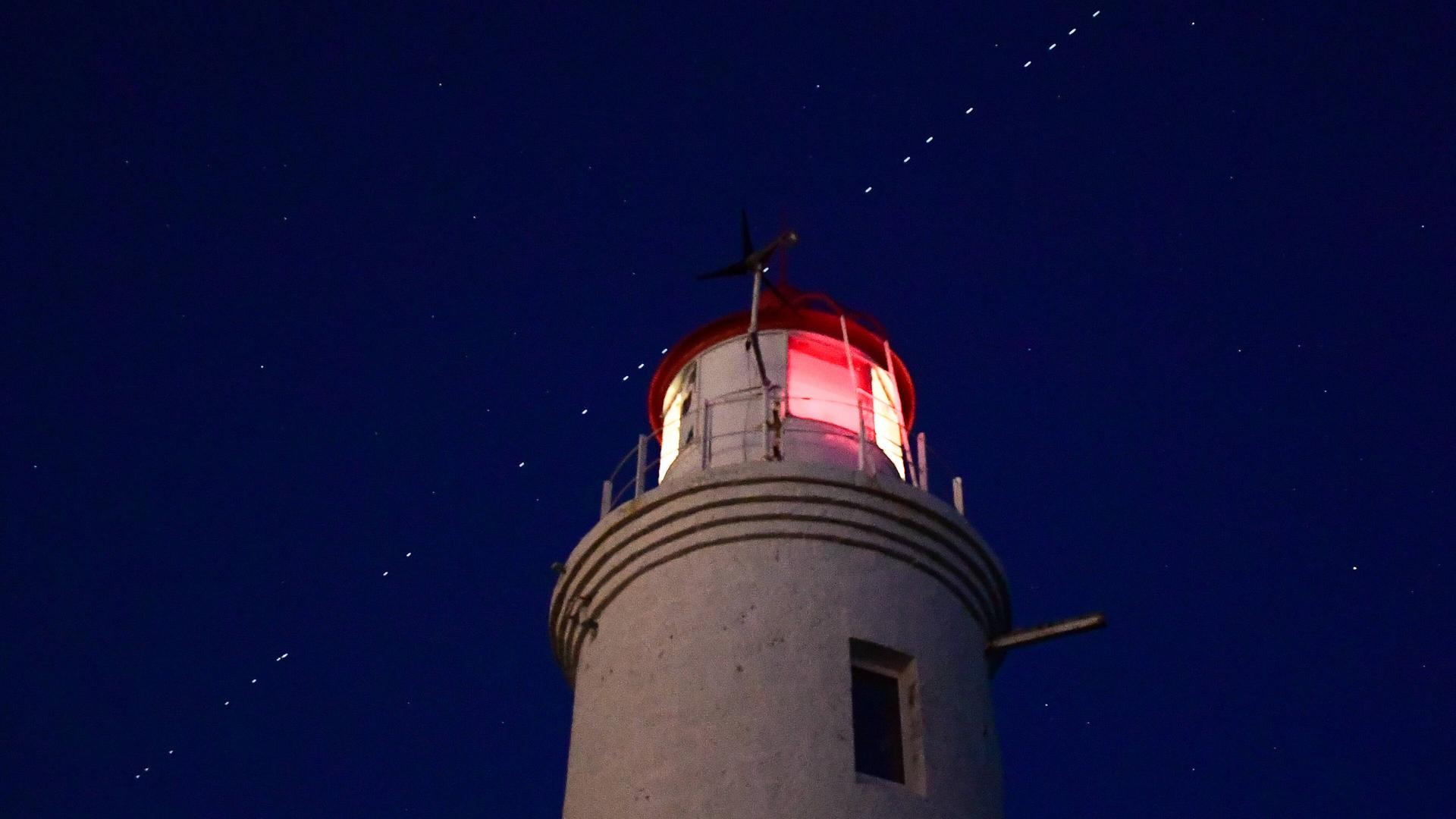 Russland, Wladiwostock: EIne Kette von Starlink Satelliten ist über einem Leuchtturm zu sehen