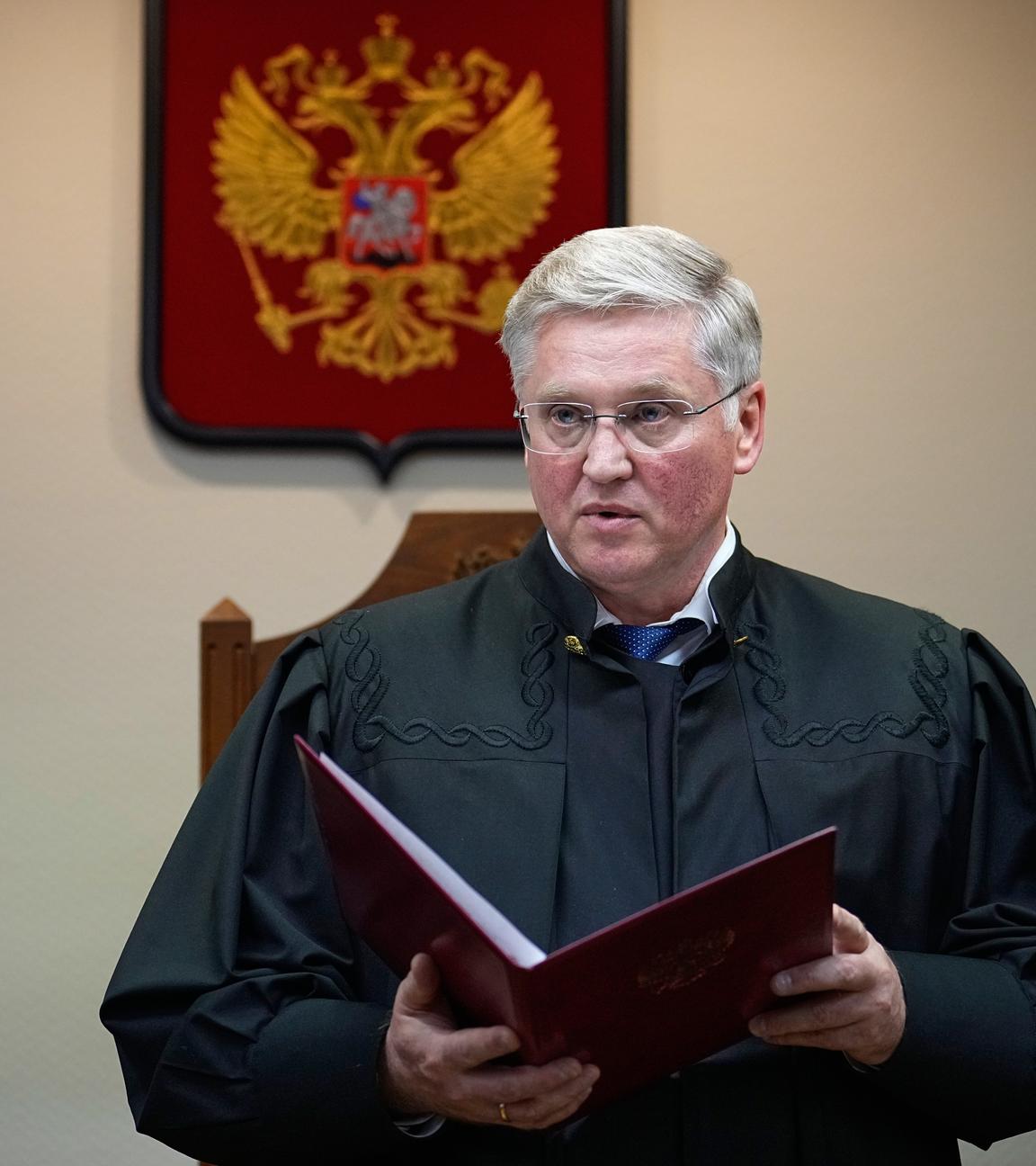 Ein Richter in einer schwarzen Robe steht in einem Gerichtssaal und hält eine große geöffnete Akte in den Händen. Hinter ihm ist das russiche Wappen zu sehen.