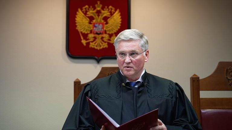 Ein Richter in einer schwarzen Robe steht in einem Gerichtssaal und hält eine große geöffnete Akte in den Händen. Hinter ihm ist das russiche Wappen zu sehen.