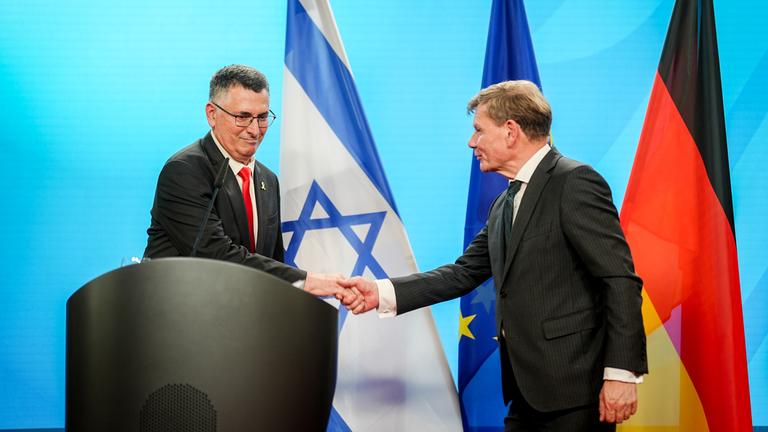 Gideon Saar und Johann Wadephul schütteln sich bei Pressekonferenz die Hände. Hinter ihnen die deutsche, israelische und EU-Flagge.
