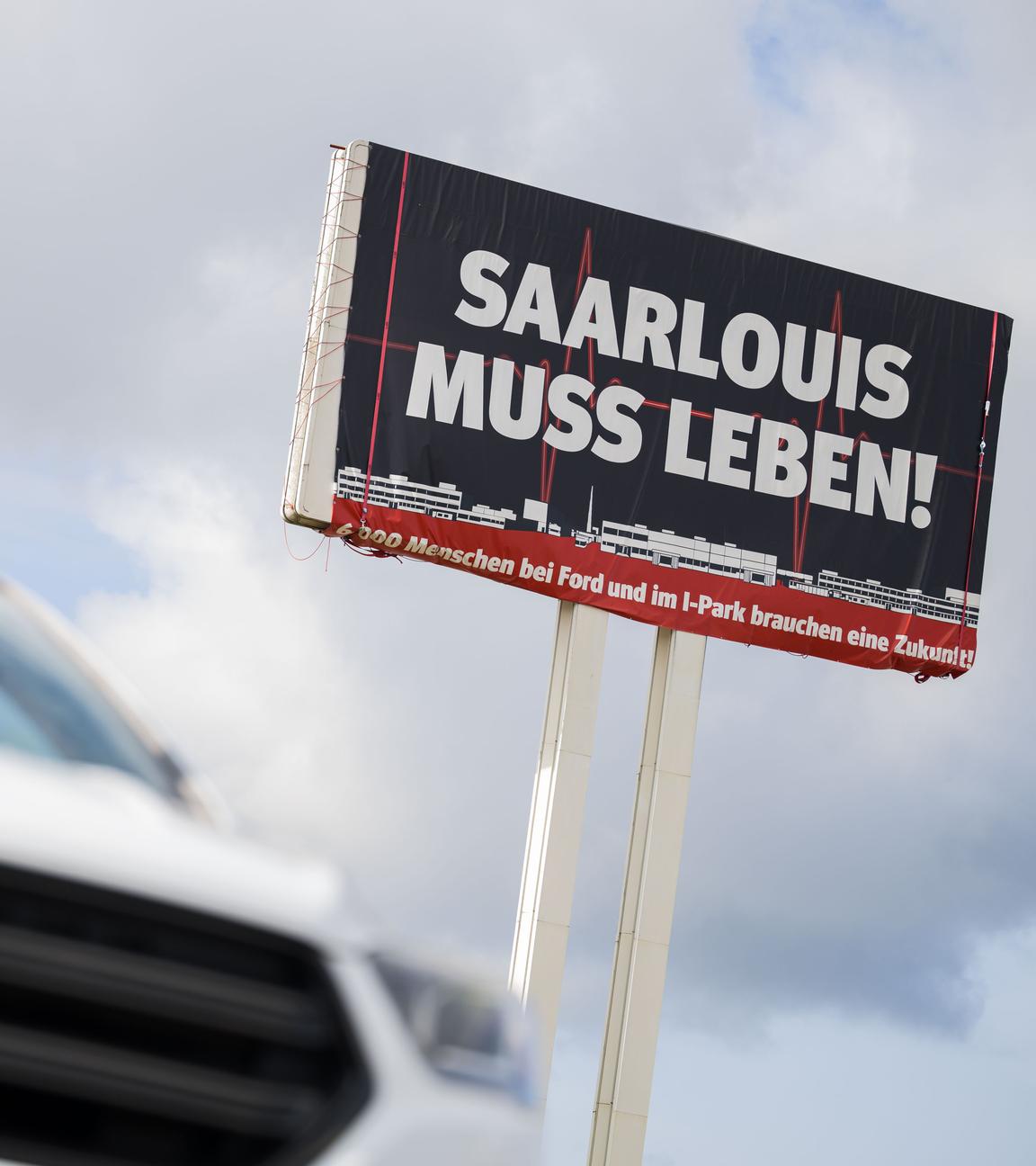 Ein Ford Kuga steht vor einem großen Schild auf dem für die Bedeutung des Ford Werks in Saarlouis geworben wird.