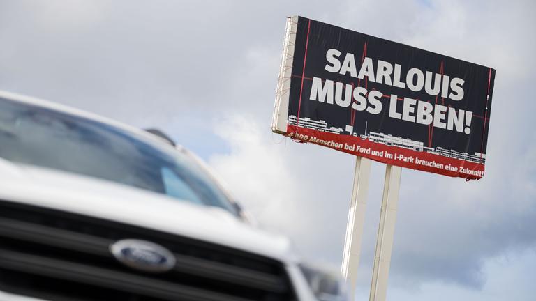 Ein Ford Kuga steht vor einem großen Schild auf dem für die Bedeutung des Ford Werks in Saarlouis geworben wird.