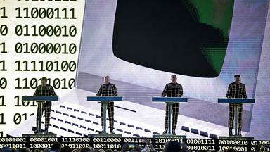 Auf dem Bild zu sehen: Vier Mitglieder der Band Kraftwerk stehen hinter elektronischen Musikpulten auf einer Bühne.