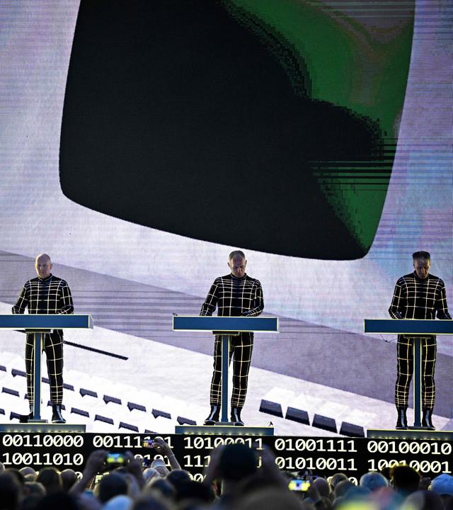 Auf dem Bild zu sehen: Vier Mitglieder der Band Kraftwerk stehen hinter elektronischen Musikpulten auf einer Bühne.