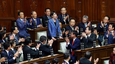 Es ist die neue Ministerpräsidentin zu sehen, wie sie im Kongress Japans steht. Um sie herum sind andere Abgeordnete.