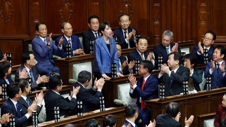 Es ist die neue Ministerpräsidentin zu sehen, wie sie im Kongress Japans steht. Um sie herum sind andere Abgeordnete.