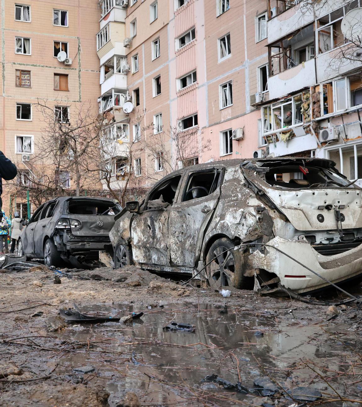 Auf der schlammbedeckten Straße vor einem Wohnblock stehen ausgebrannte Autos. Mehrere Menschen in Gummistiefeln und reflektierender Warnkleidung laufen daran vorbei.
