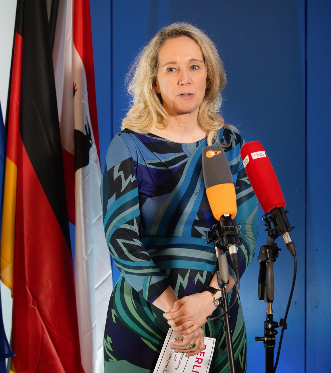 Eine Frau steht vor zwei Mikrofonen und redet. Im Hintergrund stehen Fahnen.