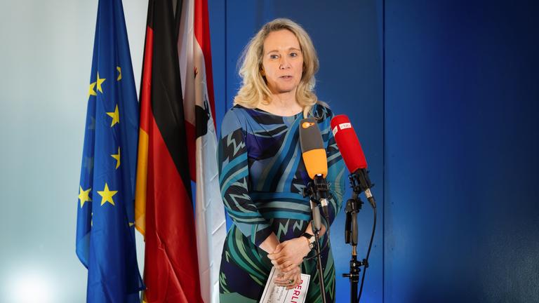 Eine Frau steht vor zwei Mikrofonen und redet. Im Hintergrund stehen Fahnen.