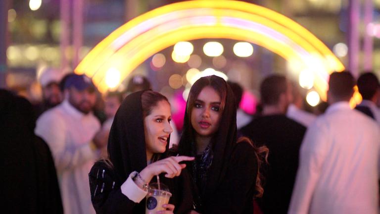 "Saudi Arabien - Öl, Tradition und Zukunft: Neue Freiheit": Abendaufnahme: Geschminkte saudische Frauen vor einer Menschenmenge. Im Hintergrund ein Lichtbogen.
