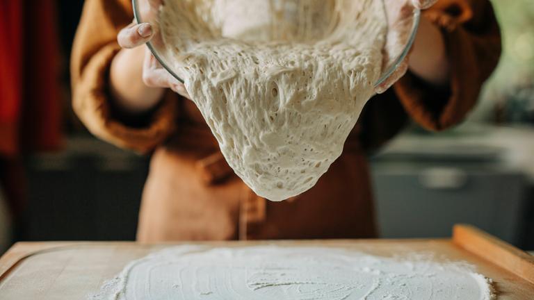 Frau lässt fermentierten Sauerteig aus einer Glasschüssel auf eine bemehlte Arbeitsfläche fließen.