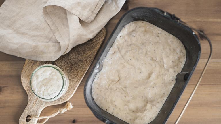 Brotteig in einer Form neben einem Brett mit einem Glas, in dem sich der Sauerteig-Ansatz befindet.