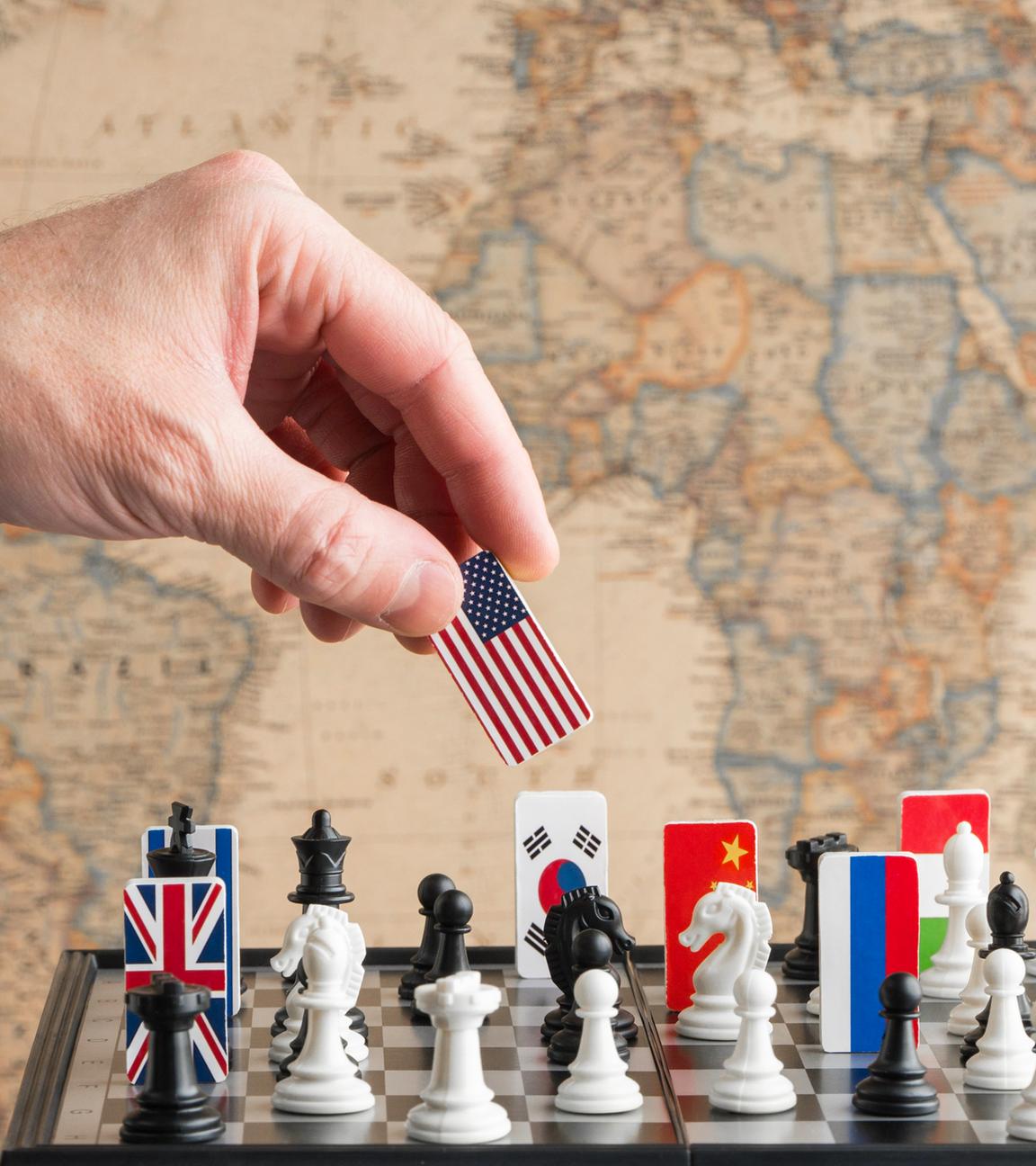 Eine Hand nimmt eine Spielfigur mit US-amerikanischer Flagge von einem Schachspiel, auf dem Länderflaggen verteilt sind.