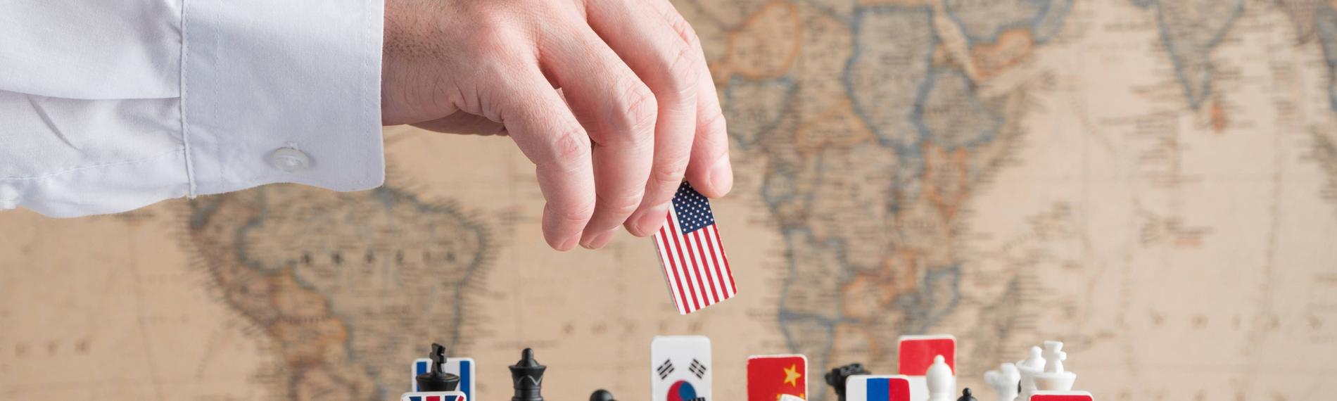 Eine Hand nimmt eine Spielfigur mit US-amerikanischer Flagge von einem Schachspiel, auf dem Länderflaggen verteilt sind.