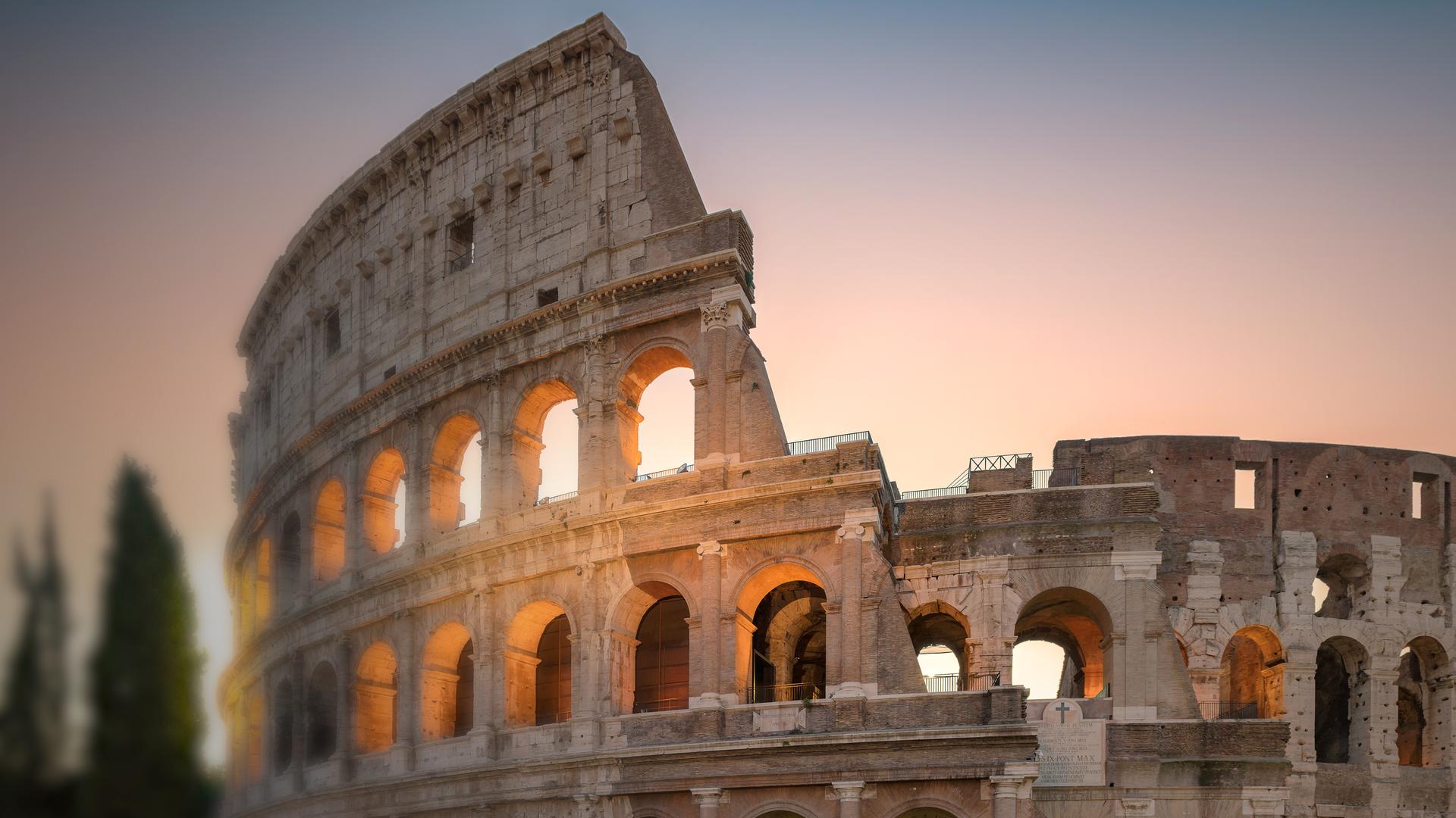 Nahaufnahme des Kolosseums in Rom von außen, der Himmel dämmert, die tiefstehende Sonne strahlt durch die Fensternischen hindurch.
