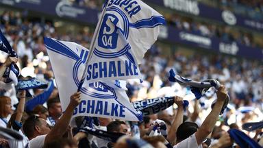 In einem Stadion steht eine Menschenmenge und hält blau-weiße Schalke Fahnen und Schals.