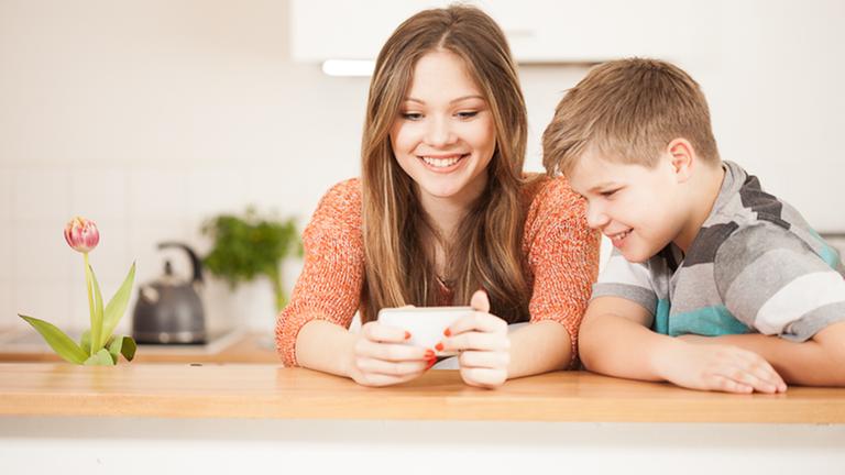 Ein bloders Mädchen im Teenie-Alter und ihr jüngerer Bruder sitzen am Tisch, schauen in ein Smartphone und lächeln.
