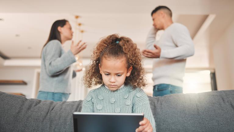 Ein Mädchen sitzt mit einem Tablet auf dem Sofa und ihre Eltern streiten sich im Hintergrund.