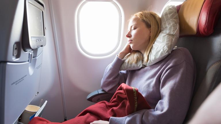 Eine Blonde Frau schläft im Flugzeug mit Nackenkissen und Decke.