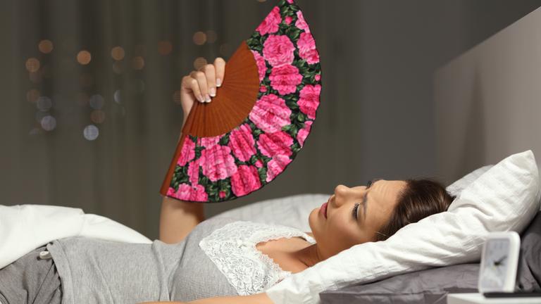 Eine Frau mit Fächer liegt im Bett und fächelt sich Luft zu.