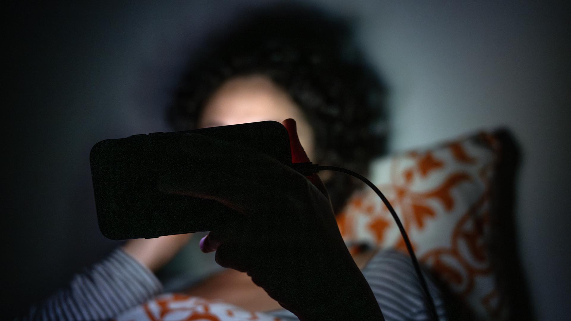 Eine Frau schaut im Bett eine Sendung auf dem Smartphone (Illustration)