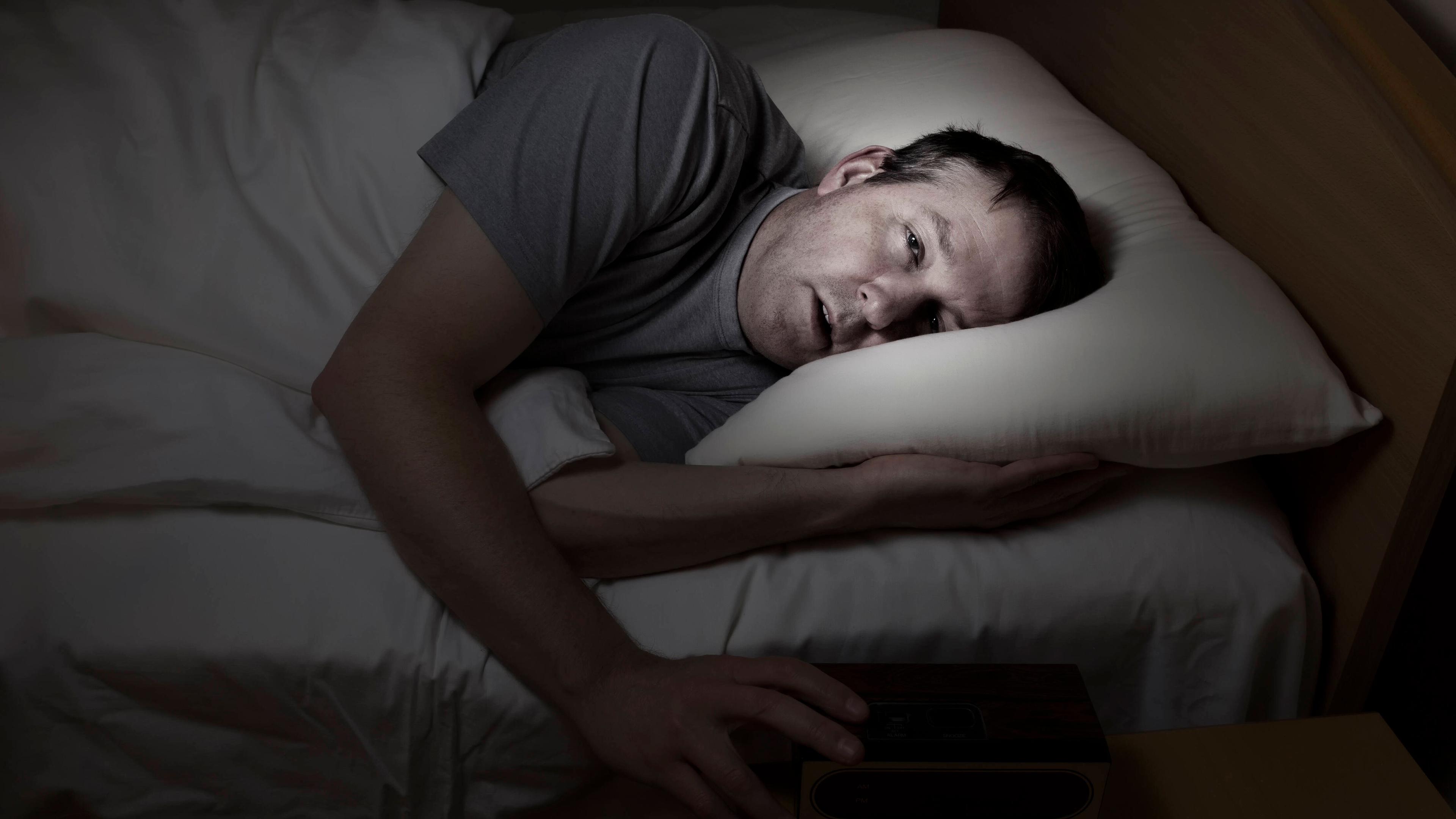 Mann liegt nachts wach im Bett mit der Hand auf dem Wecker