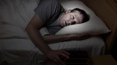 Mann liegt nachts wach im Bett mit der Hand auf dem Wecker