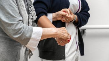 Eine Pflegerin hält eine Patientin unterstützend an den Händen.