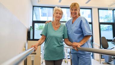 Eine ältere Frau wird von einer Krankenschwester an einer Stange gestützt.