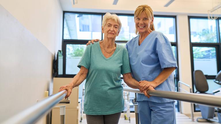 Eine ältere Frau wird von einer Krankenschwester an einer Stange gestützt.