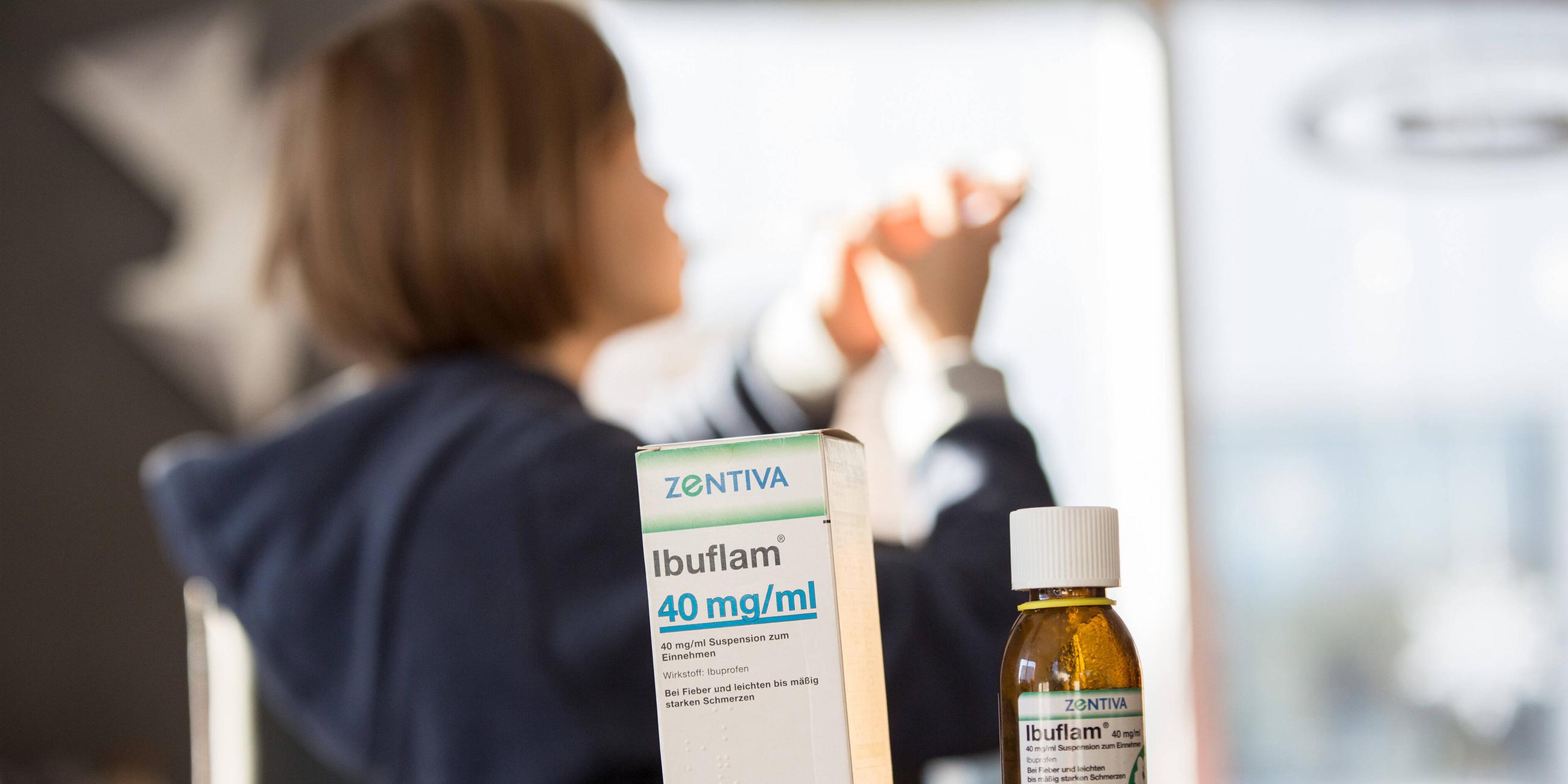 Auf einem Tisch stehen Fieber- und Schmerzmittelsaft für Kinder, im Hintergrund sitzt ein krankes Kind das den Schmerzsaft gegen Fieber sowie Schmerzen einnimmt. 