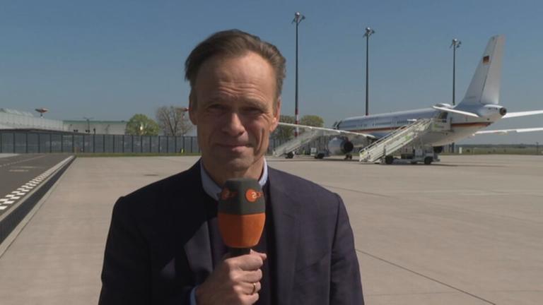 Wulf Schmiese ist mit einem Mikrofon zu sehen, im Hintergrund ist eine Fluglandebahn mit Flugzeug zu sehen.