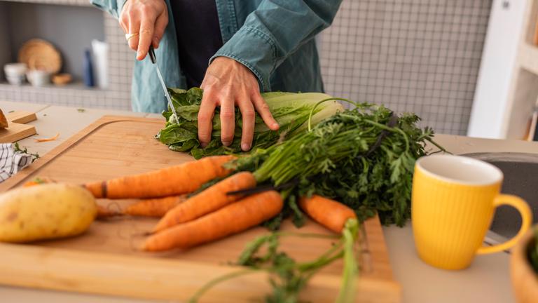 Gemüse liegt auf einem Schneidebrett aus Holz, ein Mann setzt zum Anschneiden eine Salatkopfes an.