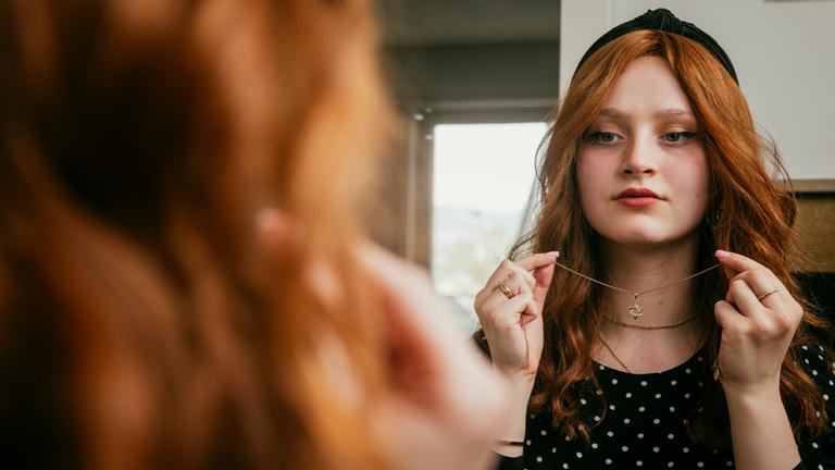 Meira steht vor einem Spiegel und legt eine Kette mit Davidstern-Anhänger an.