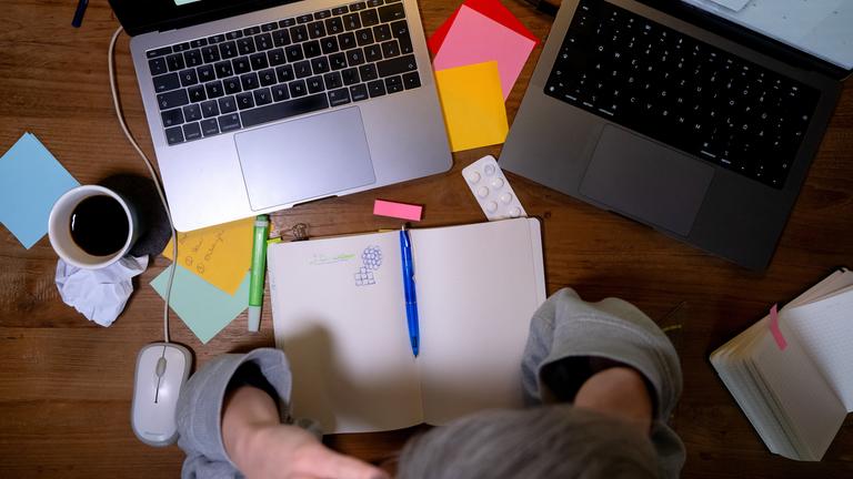 ein unordentlicher Tisch mit PCs, Zetteln und Terminkalender, eine Person hält den Kopf in den Händen