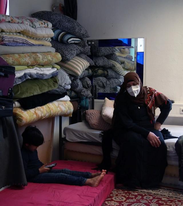 Zwei Frauen mit Hidschab (Kopftuch) sitzen in einem karg eingerichteten Zimmer auf einem Bett, daneben stapeln sich Decken und ein Kind sitzt auf einer Matratze und spielt in einem Handy