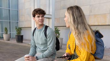 Schüler sitzen mit Rucksäcken auf einer Bank auf einem Platz und halten Kaffee und ein Smartphone in der Hand. 