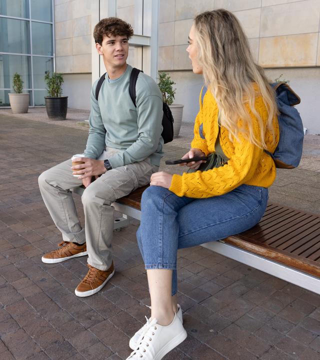 Schüler sitzen mit Rucksäcken auf einer Bank auf einem Platz und halten Kaffee und ein Smartphone in der Hand. 