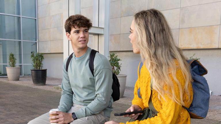 Schüler sitzen mit Rucksäcken auf einer Bank auf einem Platz und halten Kaffee und ein Smartphone in der Hand. 