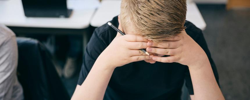 Ansicht von oben auf einen Jugendlichen, der mit dem Kopf in den Händen an einem Schreibtisch sitzt und in einem Klassenzimmer lernt.