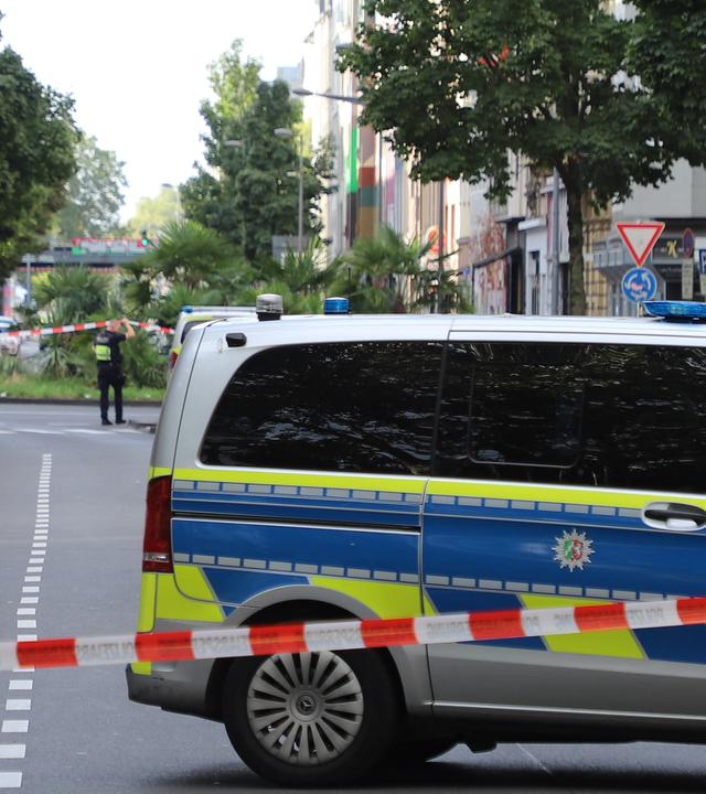 Ein Fahrzeug der Polizei steht hinter einer Absperrung auf einer Straße nachdem Schüsse am 31.08.2026 auf ein Café in Köln abgefeuert wurden.